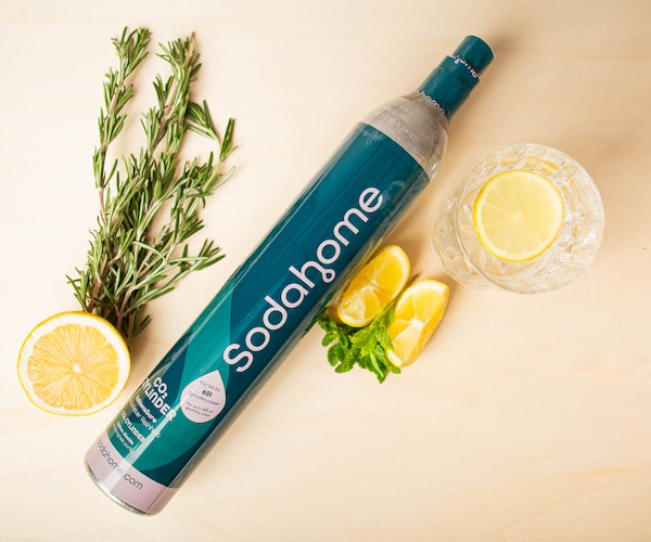Sodahome-CO2-Zylinder-Nachhaltigkeit Ein Sodahome CO₂-Zylinder liegt auf einer hellen Oberfläche, umgeben von frischen Zitronenscheiben, Kräutern und einem Glas sprudelndem Wasser mit Zitronenscheibe.