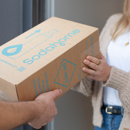 Nachhaltige Lieferung von CO₂-Zylindern mit Sodahome Übergabe eines Sodahome-Pakets an der Haustür, das umweltfreundlich verpackte CO₂-Zylinder für Wassersprudler enthält.