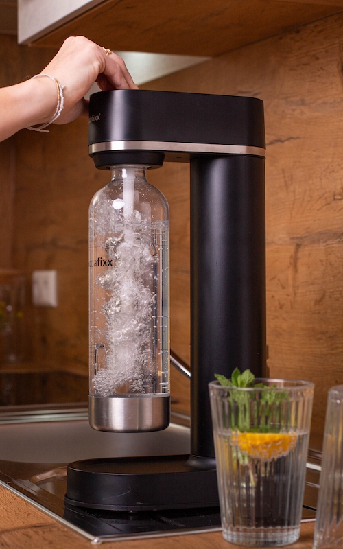 Sprudelndes Wasser mit Sodahome – Einfach und frisch Eine Hand sprudelt Wasser in einer Sodahome-Flasche mit einem schwarzen Wassersprudler. Auf der Küchenzeile stehen ein Glas mit Minze und Zitrone, frische Kräuter und eine Zitrone.