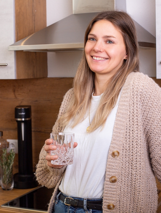 Genussvoll sprudeln mit Sodahome: Frisches Wasser auf Knopfdruck Junge Frau steht lächelnd in einer modernen Küche mit einem Glas Wasser in der Hand; im Hintergrund ein Wassersprudler und Sodahome CO₂-Zylinder.